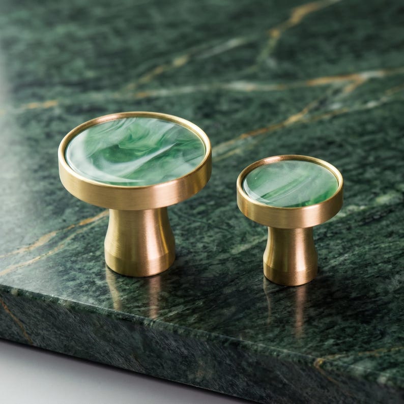 Puede incluir: Dos tiradores decorativos para armarios con bases de lat&oacute;n y tapas de m&aacute;rmol verde. Los tiradores est&aacute;n colocados sobre una superficie de m&aacute;rmol verde oscuro. Los tiradores son de diferentes tama&ntilde;os.