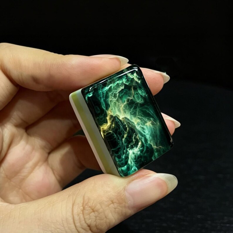 May include: A rectangular mahjong tile featuring a glossy, dark green and black surface with swirling, nebula-like patterns in shades of bright yellow and lime green. The base of the tile is a solid, opaque white material with a thin light green stripe.