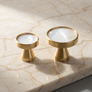 Puede incluir: Dos tiradores decorativos de lat&oacute;n con una parte superior blanca nacarada. Los tiradores tienen una base de pedestal y una parte superior circular. Los tiradores se muestran sobre una superficie de m&aacute;rmol. Ideal para muebles.