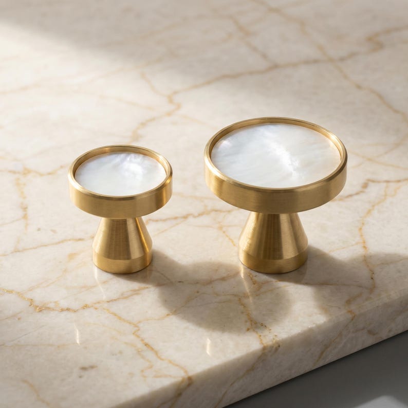 Puede incluir: Dos tiradores decorativos de lat&oacute;n con una parte superior blanca nacarada. Los tiradores tienen una base de pedestal y una parte superior circular. Los tiradores se muestran sobre una superficie de m&aacute;rmol. Ideal para muebles.