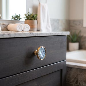 May include: A close-up of a dark gray bathroom vanity drawer with a decorative knob. The knob has a gold-colored base and a round, iridescent, blue and white center. The vanity is topped with a marble countertop.