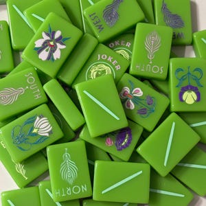 May include: A collection of bright green Mahjong tiles featuring various engraved designs including peacock feathers, floral patterns, and the words Joker, North, South, and West. Some tiles display a single diagonal line in a lighter green shade.