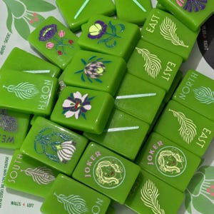 May include: A collection of bright green Mahjong tiles featuring various designs including peacock feathers, floral illustrations, and Joker characters. Some tiles are marked with the text North, South, East, West, or Joker in white lettering.