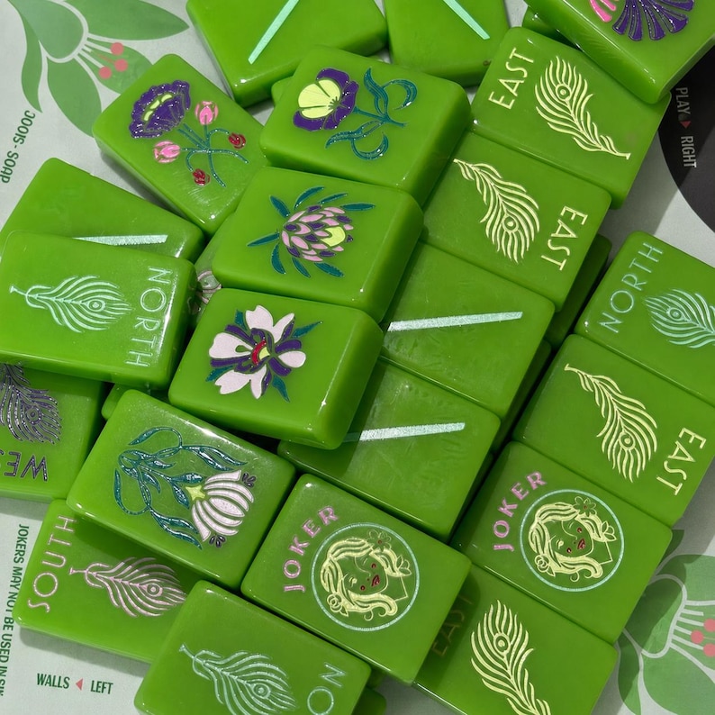 May include: A collection of bright green Mahjong tiles featuring various designs including peacock feathers, floral illustrations, and Joker characters. Some tiles are marked with the text North, South, East, West, or Joker in white lettering.