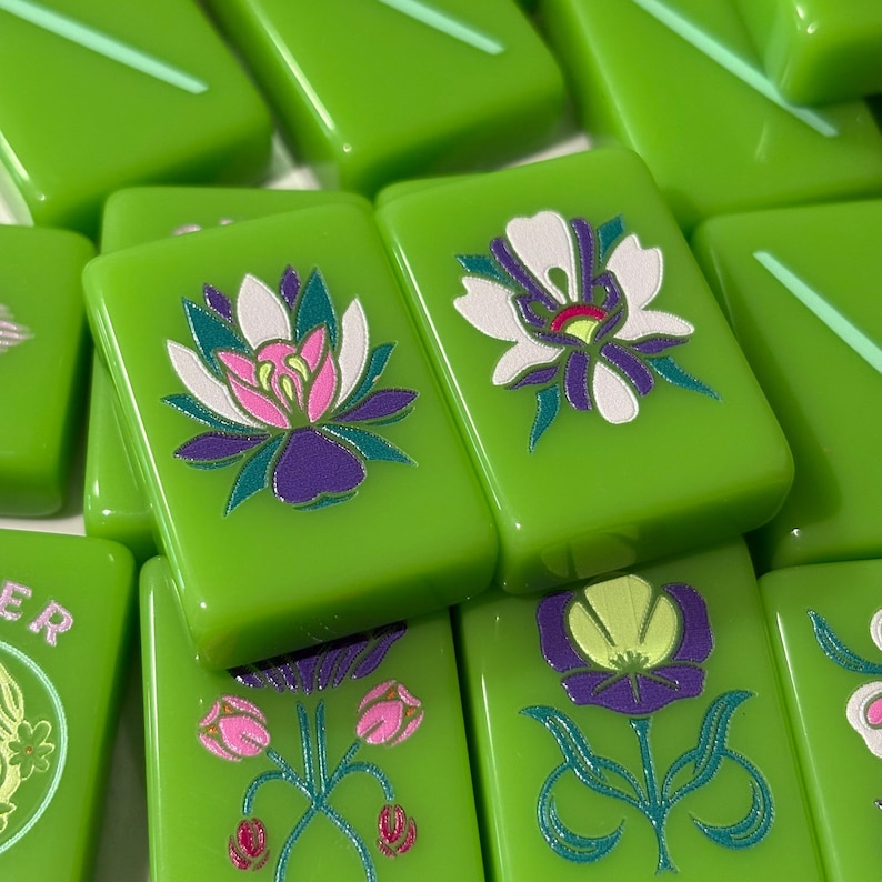 May include: A collection of rectangular green mahjong tiles featuring various floral illustrations. The tiles display detailed graphics of lotus flowers, tulips, and other blossoms in shades of white, pink, purple, and teal, with some tiles showing partial text.