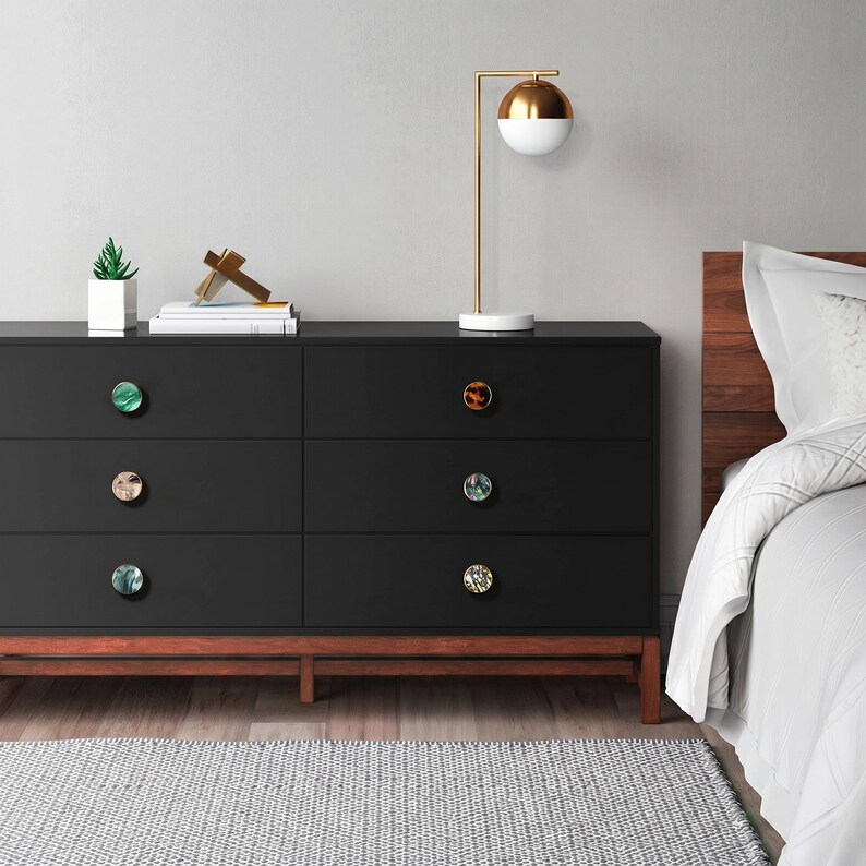 Puede incluir: Una c&oacute;moda negra de seis cajones con pomos decorativos de varios colores y dise&ntilde;os. La c&oacute;moda tiene una base de madera y est&aacute; coronada por una l&aacute;mpara dorada y blanca, una peque&ntilde;a planta y libros. Una cama con edred&oacute;n blanco est&aacute; al fondo.
