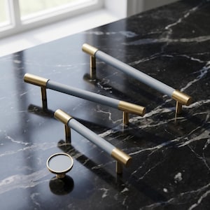 May include: A collection of cabinet hardware, including three gray and gold bar pulls and a matching round knob. The pulls have a cylindrical gray center with gold-toned ends and posts. The hardware is displayed on a black marble surface.