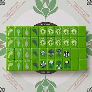 May include: A set of green Mahjong tiles arranged in a grid on a patterned surface. The tiles feature engraved designs of feathers, the words EAST, NORTH, SOUTH, WEST, the word JOKER with a face illustration, and various colorful floral motifs.