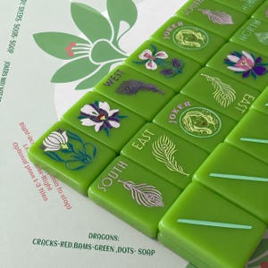 May include: A set of bright green Mahjong tiles arranged in rows, featuring engraved illustrations of flowers, peacock feathers, and Joker faces. The tiles include directional labels like North, South, East, and West, with text detailing game rules.