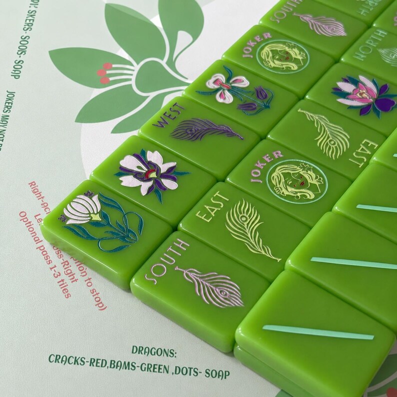 May include: A set of bright green Mahjong tiles arranged in rows, featuring engraved illustrations of flowers, peacock feathers, and Joker faces. The tiles include directional labels like North, South, East, and West, with text detailing game rules.