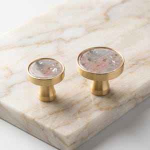 Puede incluir: Dos tiradores de armario con acabado dorado y una parte superior redonda y transparente con un dise&ntilde;o de piedra rosa y gris. Los tiradores est&aacute;n sobre una superficie de m&aacute;rmol. Estos tiradores decorativos son ideales para a&ntilde;adir un toque de elegancia a los muebles.