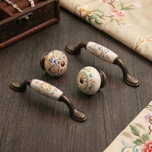 May include: A collection of decorative cabinet hardware, including two knobs and two pulls. The knobs and pulls feature a white ceramic base with hand-painted floral designs in blue, yellow, and green. The hardware has a bronze finish.