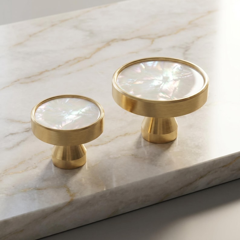 Puede incluir: Dos tiradores decorativos para armarios con acabado dorado e incrustaci&oacute;n redonda e iridiscente de n&aacute;car. Los tiradores son de diferentes tama&ntilde;os y est&aacute;n sobre una superficie de m&aacute;rmol. Ideales para a&ntilde;adir elegancia a los muebles.