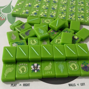 May include: A set of bright green mahjong tiles featuring various illustrations including peacock feathers, floral designs, joker faces, and directional labels like North, South, East, and West. The tiles are arranged in rows and scattered piles.