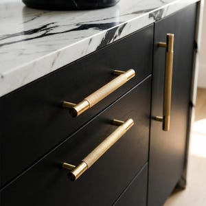 May include: Close-up of a black cabinet with gold-toned hardware. The cabinet has a marble countertop. The hardware includes two horizontal bar pulls and one vertical pull, all with a textured design.