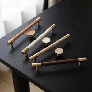 May include: A collection of cabinet hardware, including brass-toned pulls with tan, white, and brown leather-wrapped handles. Coordinating round knobs with brass bases and white or tan tops are also displayed on a black surface.