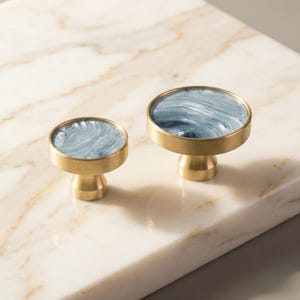 Puede incluir: Dos tiradores decorativos para armarios. Cada tirador presenta un dise&ntilde;o redondo veteado en azul y blanco, enmarcado en un metal dorado. Los tiradores tienen una base corta y cil&iacute;ndrica, y se muestran sobre una superficie de m&aacute;rmol.