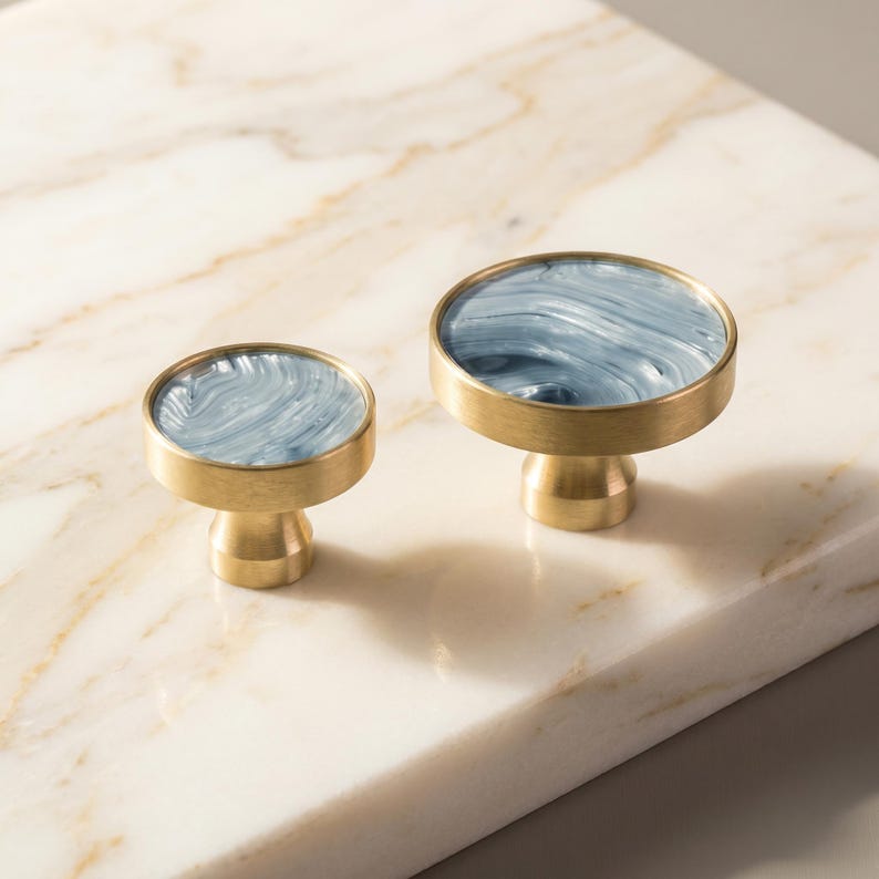 Puede incluir: Dos tiradores decorativos para armarios. Cada tirador presenta un dise&ntilde;o redondo veteado en azul y blanco, enmarcado en un metal dorado. Los tiradores tienen una base corta y cil&iacute;ndrica, y se muestran sobre una superficie de m&aacute;rmol.