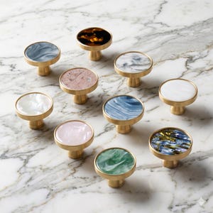 Puede incluir: Una colección de diez tiradores decorativos para armarios con bases de latón y tapas de varios colores. Las tapas presentan diseños en tonos de azul, rosa, blanco, verde y carey. Los tiradores están dispuestos sobre una superficie de mármol.