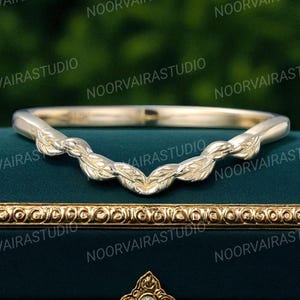 Peut inclure: Une bague dorée ornée d'un motif de feuilles. La bague a une forme incurvée avec des motifs de feuilles détaillés. Elle est présentée sur une surface vert foncé. Le nom de la marque "NOORVAIRASTUDIO" est répété en arrière-plan.