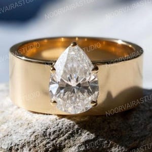 Pode incluir: Um anel de ouro com um diamante em forma de pêra. O anel tem uma banda larga e plana. O diamante é realçado por uma cravação simples. O anel é exibido em uma pedra cinza texturizada.