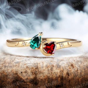 May include: A gold ring featuring a pear-shaped green gemstone and a heart-shaped red gemstone, accented by small clear stones and crescent moon engravings on the band. The design showcases two distinct gemstone cuts set in a polished gold metal band.