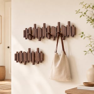 Puede incluir: Dos percheros de pared de madera marrón oscuro con un diseño de listones verticales escalonados. El perchero más grande cuenta con varios ganchos extensibles, uno de los cuales sostiene un bolso de lona beige con correas de cuero marrón.