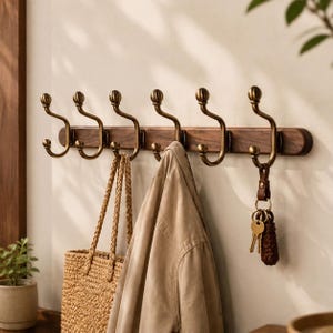 Può includere: Un appendiabiti da parete con una base orizzontale in legno scuro e sei ganci doppi in ottone anticato. I ganci presentano terminali arrotondati e bracci curvi. Una giacca beige e una borsa in paglia intrecciata sono appese ai ganci con chiavi.