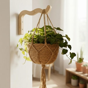 Może przedstawiać: Wiszący kosz na rośliny wykonany z plecionego włókna naturalnego, zawieszony na zakrzywionym drewnianym wsporniku. Kosz zawiera bujną zieloną roślinę z opadającymi pędami. Projekt łączy style rustykalne i nowoczesne.