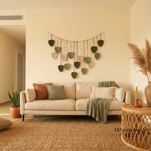 May include: A wall hanging featuring a series of suspended heart-shaped leaves in shades of sage green and cream, arranged in a cascading arc. The leaves are attached to a horizontal cord, creating a layered, symmetrical display on the wall.