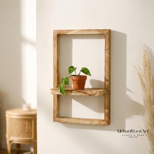 Op de afbeelding: Een rechthoekige wandplank van lichtbruin hout met één horizontale plank. Een kleine terracotta pot met een groene bladplant staat op de plank. De tekst UrbanRootArt PLANTS & HOME DECOR staat in de rechterbenedenhoek gedrukt.