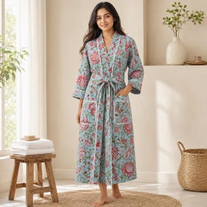 May include: A light blue robe featuring a repeating floral pattern in shades of red and green. The robe has long sleeves, a tie waist, and two front pockets, all accented with white piping along the edges, collar, and pocket openings.
