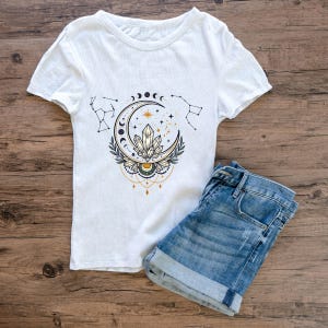Puede incluir: Camiseta blanca de manga corta con un diseño celestial que presenta una luna creciente, cristales, estrellas y constelaciones. Combinada con pantalones cortos de mezclilla azules, la camisa tiene una textura acanalada.