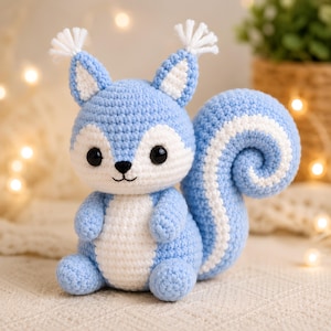 May include: A crocheted squirrel figurine crafted from light blue and white yarn. The squirrel features large black safety eyes, a small black embroidered nose and mouth, pointed ears with white tufts, and a large, curled, striped tail.