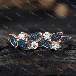 May include: A rose gold ring featuring a leaf-like design with dark blue opal stones and clear round accent stones. The opal stones have a colorful, iridescent appearance. The ring is set against a dark wooden surface.