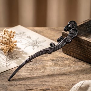 May include: A dark brown wooden hair stick featuring a smooth, tapered shaft and an ornate, curved head with carved, wave-like swirling patterns. The decorative top section displays intricate, raised scrollwork that tapers into a pointed end.