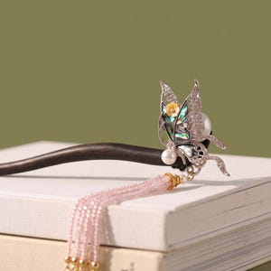 May include: A dark wooden hair stick featuring a silver-toned butterfly ornament with iridescent abalone shell inlay, a small yellow flower accent, and two white pearls. A long, delicate tassel made of light pink beads hangs from the butterfly.