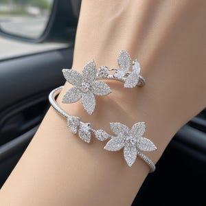 May include: A silver-toned bypass bracelet featuring two floral motifs encrusted with clear, sparkling stones. The flexible, coiled band is textured with a fine, ribbed pattern, and the flowers are accented with additional marquise-cut stones.