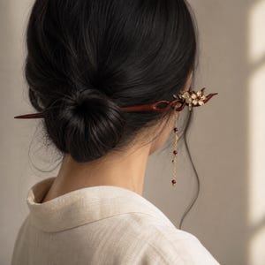 May include: A dark brown wooden hair stick featuring a carved twisted design and a decorative floral accent with small white petals. A delicate gold-toned chain hangs from the floral piece, adorned with tiny dark red beads that dangle downward.