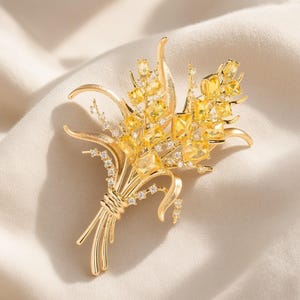May include: A gold-toned metal brooch shaped like a bouquet of wheat, featuring multiple rectangular yellow crystals and small clear accent stones. The design includes curved metal stems tied together at the base with a wrapped wire detail.