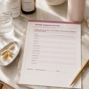 Könnte beinhalten: Ein Monthly Symptoms Overview Tracking-Blatt mit rosa Kopfzeile, das ein Raster zur täglichen Symptomerfassung wie Hitzewallungen und Stimmungsschwankungen enthält, begleitet von einem goldenen Stift auf einer weißen Marmoroberfläche mit Nahrungsergänzungsmitteln.