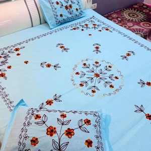 Puede incluir: Un juego de cama azul claro con diseños florales bordados. La ropa de cama presenta flores naranjas y marrones con tallos negros y un borde decorativo. El juego está diseñado para una cama.