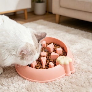 Puede incluir: Un cuenco rosa claro para comida de gato con un diseño de alimentación lenta. El cuenco está lleno de comida seca para gatos y tiene varias formas geométricas rosas en su interior. Un gato blanco está comiendo del cuenco.