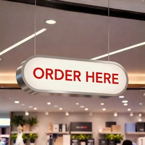 Pode incluir: Uma placa retangular iluminada com bordas arredondadas e moldura prateada. Exibe as palavras «ORDER HERE» em vermelho sobre um fundo branco. A placa está suspensa em um ambiente comercial, provavelmente uma loja.