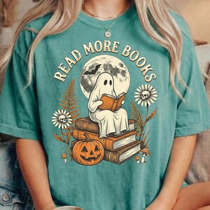 Könnte beinhalten: Ein kurzärmeliges T-Shirt in Petrol mit einer Grafik eines Geistes, der auf einem Bücherstapel sitzt und liest, umgeben von einem Kürbis, Blumen und einem Vollmond. Der Text READ MORE BOOKS ist in einem Bogen über dem Motiv gedruckt.
