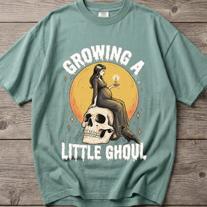 May include: A light green t-shirt featuring a graphic print of a pregnant figure sitting on a large skull in front of a yellow moon. The figure holds a lit candle, and a black crow stands nearby. The text reads GROWING A LITTLE GHOUL in white.