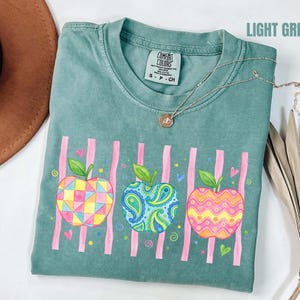 Może przedstawiać: Jasnozielony t-shirt z grafiką przedstawiającą trzy jabłka o różnych wzorach: jedno w kolorowe figury geometryczne, drugie w niebieski wzór paisley, a trzecie w pomarańczowe faliste linie, na tle pionowych różowych pasków i małych serduszek.