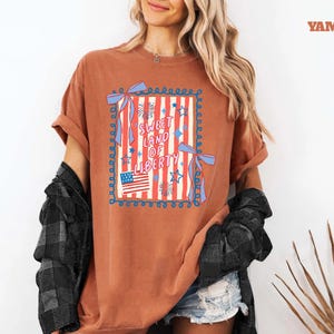 May include: A rust-colored t-shirt with a patriotic design. The graphic features the text "Sweet Land of Liberty" in a square frame with red and white stripes, stars, and an American flag. The shirt has rolled-up sleeves.