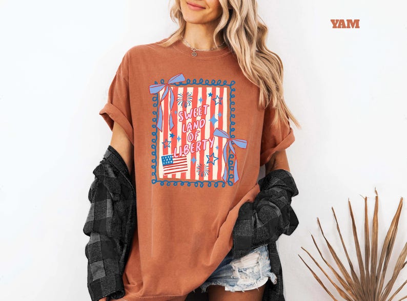 May include: A rust-colored t-shirt with a patriotic design. The graphic features the text "Sweet Land of Liberty" in a square frame with red and white stripes, stars, and an American flag. The shirt has rolled-up sleeves.