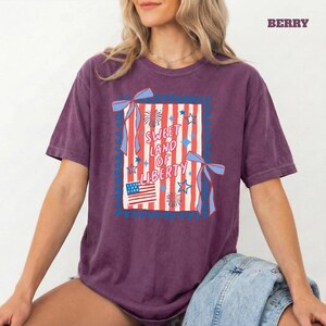 May include: Berry-colored t-shirt with a patriotic design. The graphic features red and white stripes, blue ribbons, stars, and the text "Sweet Land of Liberty." An American flag is also included in the design.